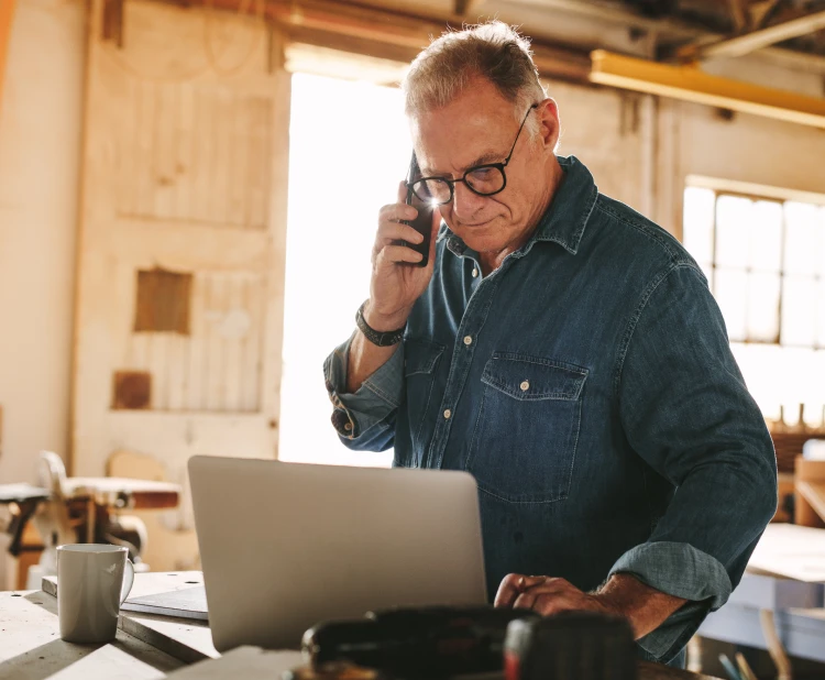 An old man is talking with customers on his mobile and browsing his laptop.