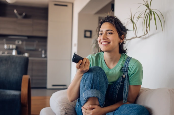 A young woman sitting on the couch and smiling while pointing a remote control to the tv.