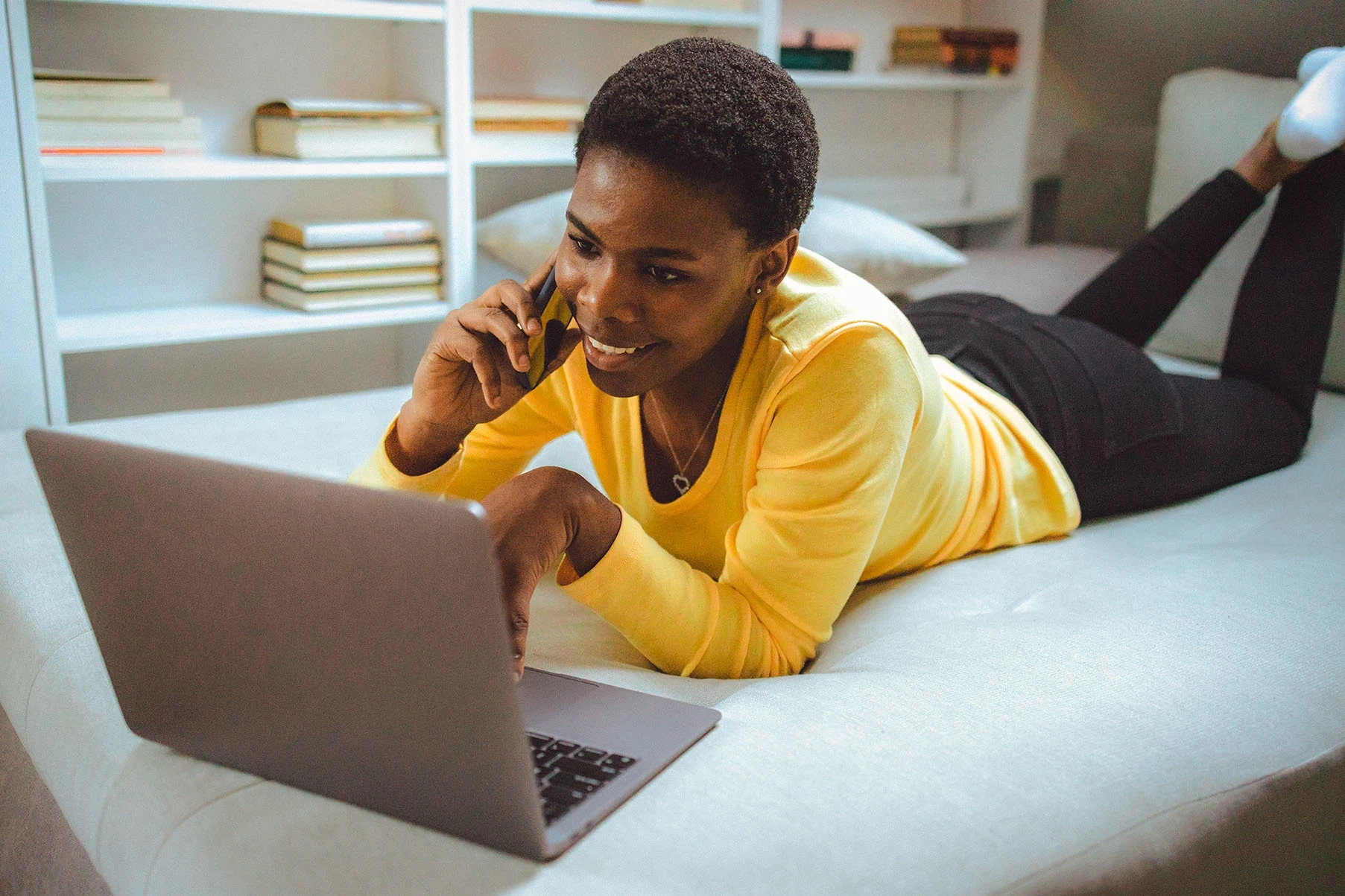 Woman lying on her stomach on her phone with laptop in front of her